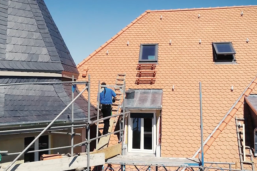 Dachdecker steht auf einem Gerüst vor einem Dach, welches gerade mit Ziegeln belegt wird.